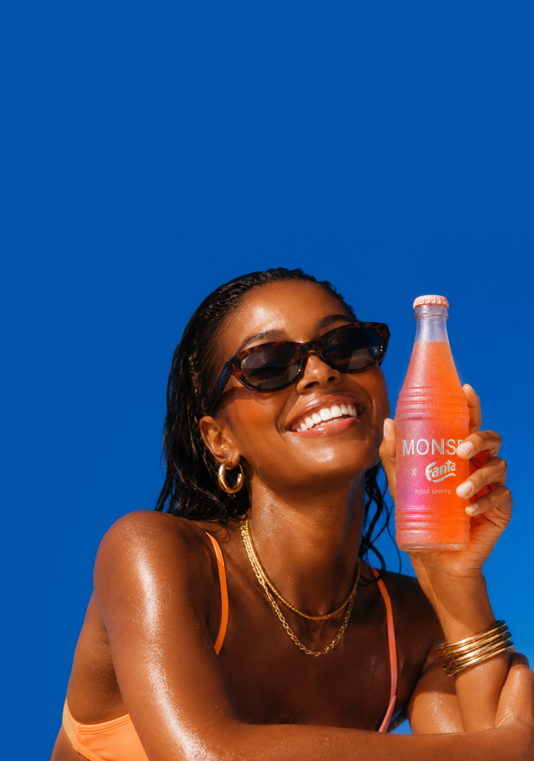 A woman in the sun holding a chilled MONSE × Fanta Rosé Spritz bottle against a deep blue sky